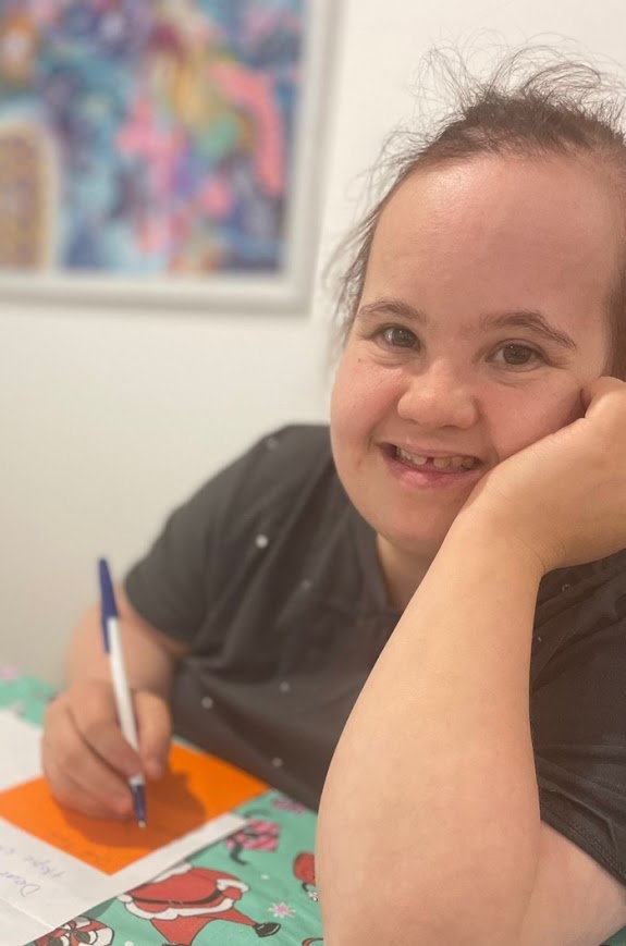 Emily smiles while writing with a pen at a table, her head resting on her hand. She is wearing a dark shirt and sitting in a brightly lit room with colorful artwork in the background.