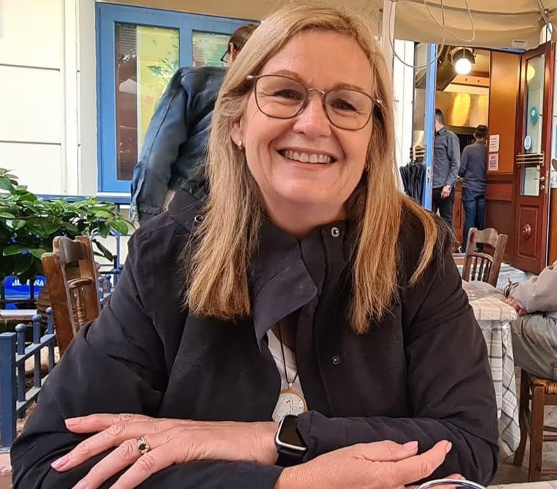 Cassie with blonde hair and glasses smiles while sitting at a table in an outdoor café. She is wearing a dark jacket, and there are people and a colorful storefront in the background.