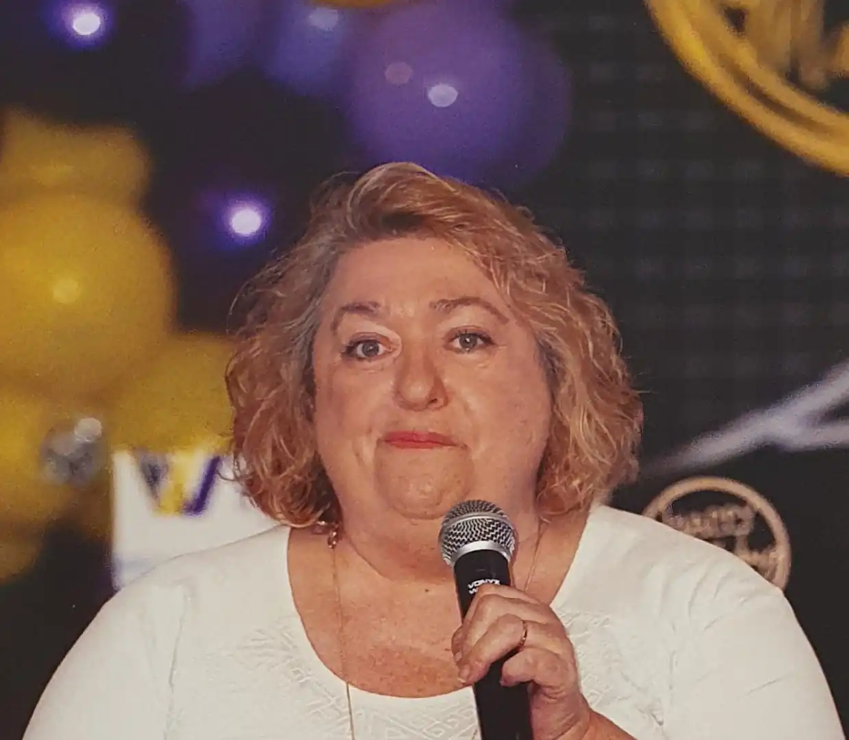 Jenny with curly blonde hair wearing a white top, holding a microphone and speaking at an event with a background of purple and yellow balloons.