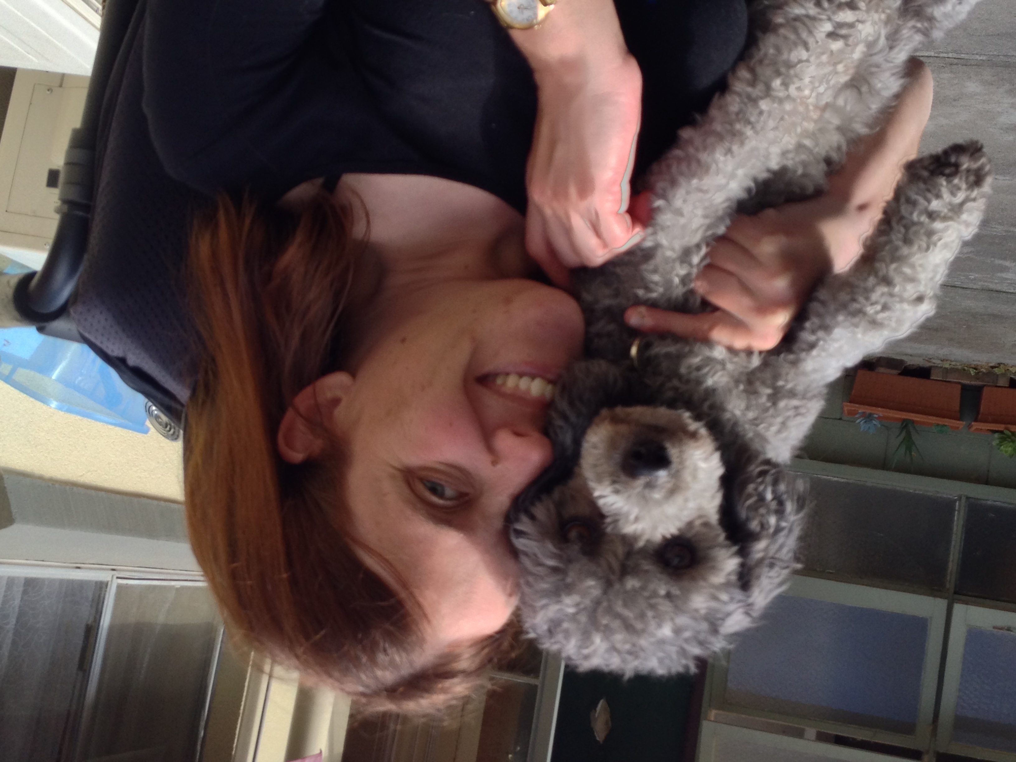 Kate with brown hair smiles while holding a gray poodle in a patio area with potted plants and windows in the background.