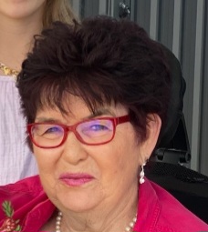 Liz with short dark hair and red glasses smiles while wearing a red top and pearl earrings. She is in an indoor setting with part of another person visible behind her.