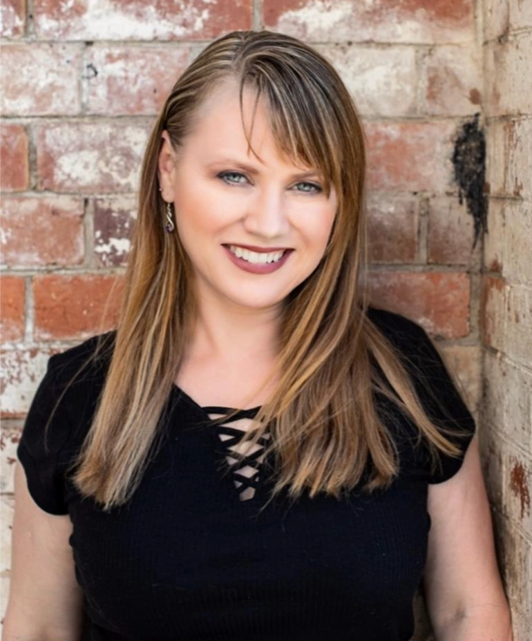 Rachel with straight, light brown hair and side-swept bangs smiles while standing in front of a brick wall. She is wearing a black top with a crisscross design at the neckline.
