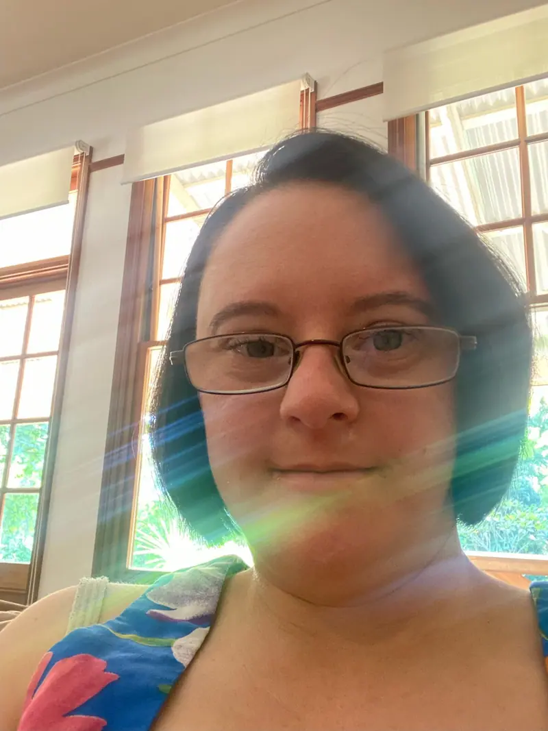 Ruth with short dark hair and glasses takes a selfie indoors. She is wearing a colorful top, and sunlight is streaming through the windows behind her, creating a lens flare effect.